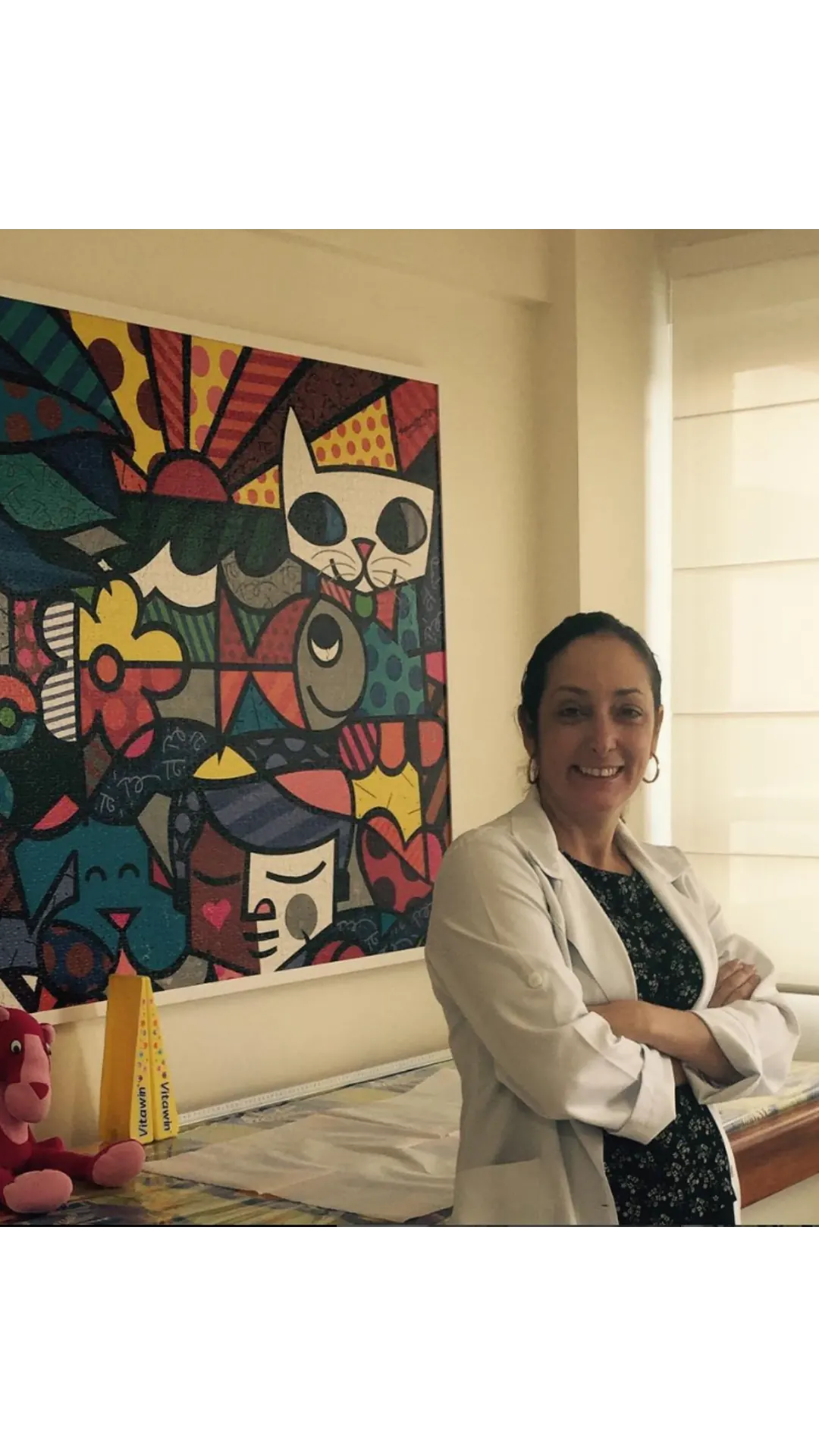 Dra. Sabrina Nunes de jaleco no consultório, ao lado de um quadro colorido.