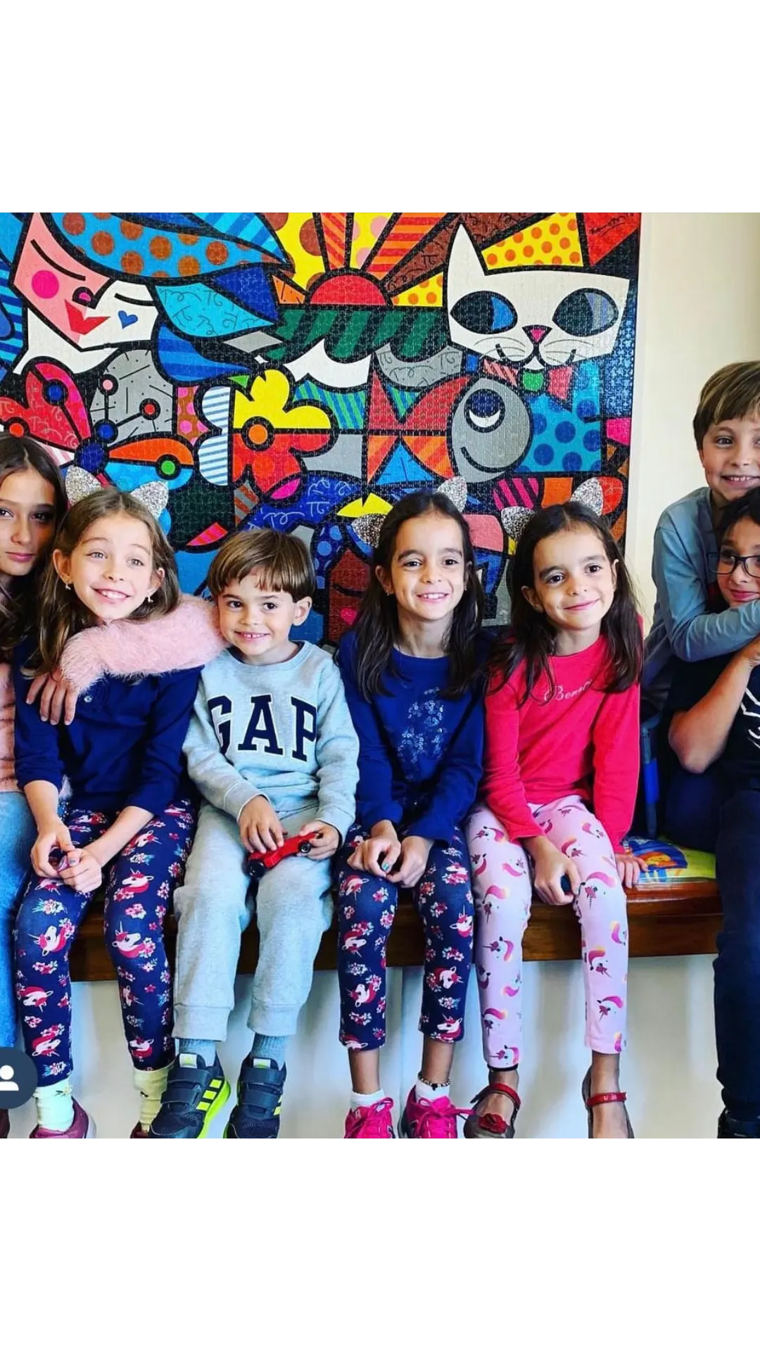 Grupo de crianças sorrindo, sentadas em um banco em frente a um quadro colorido.